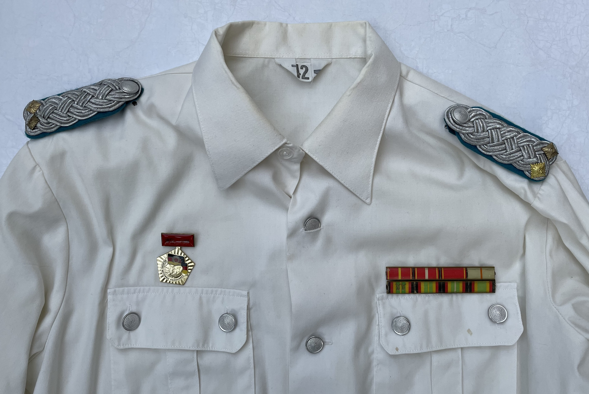 En vit skjortblus (Hemdbluse) för överstelöjtnant vid de östtyska gränstrupperna (Grenztruppen der DDR). Storlek 42N. Skjortblusen ingick i stabsuniform sommar (Stabsdienstuniform Nr. 2 So) och bars utan skjorta under.
Över höger bröstficka sitter en minnesmedalj för östtyska armens (Nationalen Volksarmee – NVA) 30-års jubileum. På nålfästet texten ”1956-1986”, på medaljens framsida ”30 JAHRE NATIONALE VOLKSARMEE” och på frånsidan ”Fest verwurzelt im Volk, gefürt von der Partei, erfüllen wir den Klassenauftrag. Forwärts zom XI. Parteitag der SEDI”.
Över vänster bröstficka sitter släpspännen. 
I övre raden band för (från höger till vänster från betraktarens håll); Vapenbrödramedaljen (Medaille der Waffenbrüderschaft) i brons och östtyska armens förtjänstmedalj (Verdienstmedaille der Nationalen Volksarmee) i brons, silver och guld.
I undre raden band för (från höger till vänster från betraktarens håll); medaljen för trogen tjänst i östtyska gränstrupperna (Medaille für treue Dienste in den Grenztruppen der DDR) 5 års tjänst, 10 års tjänst, 15 års tjänst och 20 års tjänst.