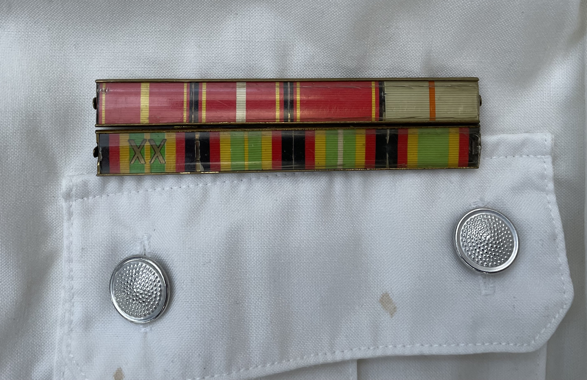 En vit skjortblus (Hemdbluse) för överstelöjtnant vid de östtyska gränstrupperna (Grenztruppen der DDR). Storlek 42N. Skjortblusen ingick i stabsuniform sommar (Stabsdienstuniform Nr. 2 So) och bars utan skjorta under.
Över höger bröstficka sitter en minnesmedalj för östtyska armens (Nationalen Volksarmee – NVA) 30-års jubileum. På nålfästet texten ”1956-1986”, på medaljens framsida ”30 JAHRE NATIONALE VOLKSARMEE” och på frånsidan ”Fest verwurzelt im Volk, gefürt von der Partei, erfüllen wir den Klassenauftrag. Forwärts zom XI. Parteitag der SEDI”.
Över vänster bröstficka sitter släpspännen. 
I övre raden band för (från höger till vänster från betraktarens håll); Vapenbrödramedaljen (Medaille der Waffenbrüderschaft) i brons och östtyska armens förtjänstmedalj (Verdienstmedaille der Nationalen Volksarmee) i brons, silver och guld.
I undre raden band för (från höger till vänster från betraktarens håll); medaljen för trogen tjänst i östtyska gränstrupperna (Medaille für treue Dienste in den Grenztruppen der DDR) 5 års tjänst, 10 års tjänst, 15 års tjänst och 20 års tjänst.