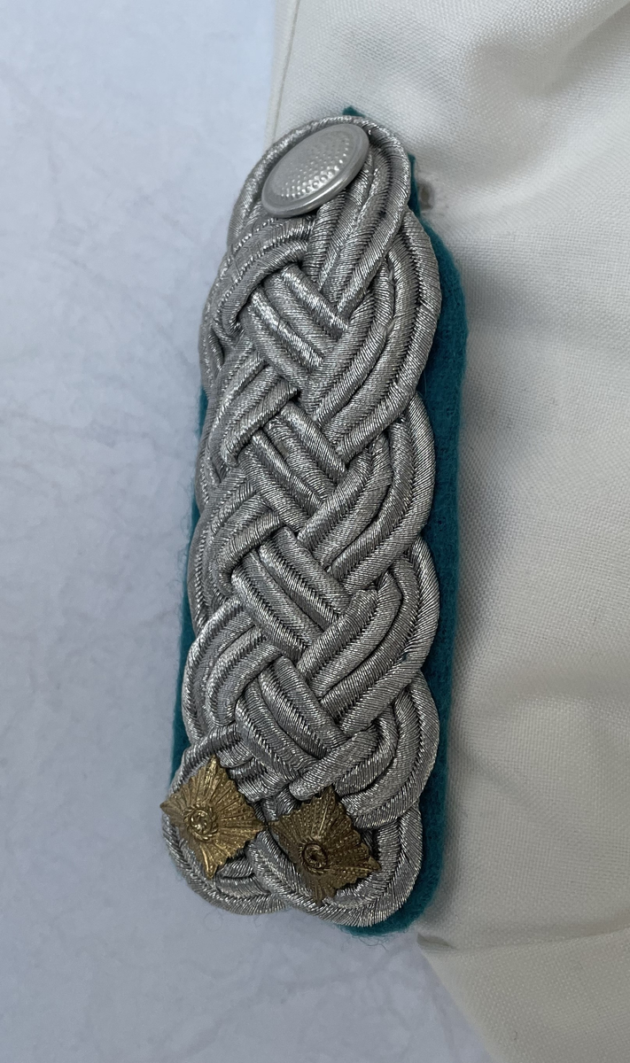 En vit skjortblus (Hemdbluse) för överstelöjtnant vid de östtyska gränstrupperna (Grenztruppen der DDR). Storlek 42N. Skjortblusen ingick i stabsuniform sommar (Stabsdienstuniform Nr. 2 So) och bars utan skjorta under.
Över höger bröstficka sitter en minnesmedalj för östtyska armens (Nationalen Volksarmee – NVA) 30-års jubileum. På nålfästet texten ”1956-1986”, på medaljens framsida ”30 JAHRE NATIONALE VOLKSARMEE” och på frånsidan ”Fest verwurzelt im Volk, gefürt von der Partei, erfüllen wir den Klassenauftrag. Forwärts zom XI. Parteitag der SEDI”.
Över vänster bröstficka sitter släpspännen. 
I övre raden band för (från höger till vänster från betraktarens håll); Vapenbrödramedaljen (Medaille der Waffenbrüderschaft) i brons och östtyska armens förtjänstmedalj (Verdienstmedaille der Nationalen Volksarmee) i brons, silver och guld.
I undre raden band för (från höger till vänster från betraktarens håll); medaljen för trogen tjänst i östtyska gränstrupperna (Medaille für treue Dienste in den Grenztruppen der DDR) 5 års tjänst, 10 års tjänst, 15 års tjänst och 20 års tjänst.