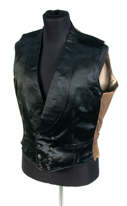 Väst av svart siden med stora slag, dubbelknäppt, 4 flata, små sidenöverklädda knappar i var rad. Två raka fickor. Gult linnefoder. I kanten och på ryggens baksida, brunt linnelärft. (På vänstra sidan nedtill ligger insytt 1 hålsmatt, hårt föremål.