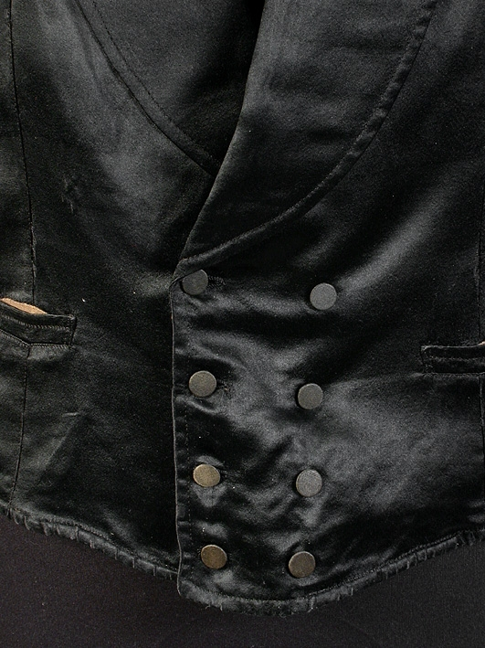 Väst av svart siden med stora slag, dubbelknäppt, 4 flata, små sidenöverklädda knappar i var rad. Två raka fickor. Gult linnefoder. I kanten och på ryggens baksida, brunt linnelärft. (På vänstra sidan nedtill ligger insytt 1 hålsmatt, hårt föremål.