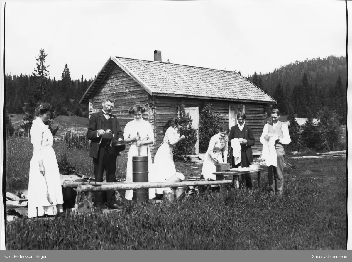 Fäbodbild från Klackbodarna i Stöde. Framför en fäbodstuga poserar fyra kvinnor och tre män med smörkärna, kaffepetter och diskbalja.