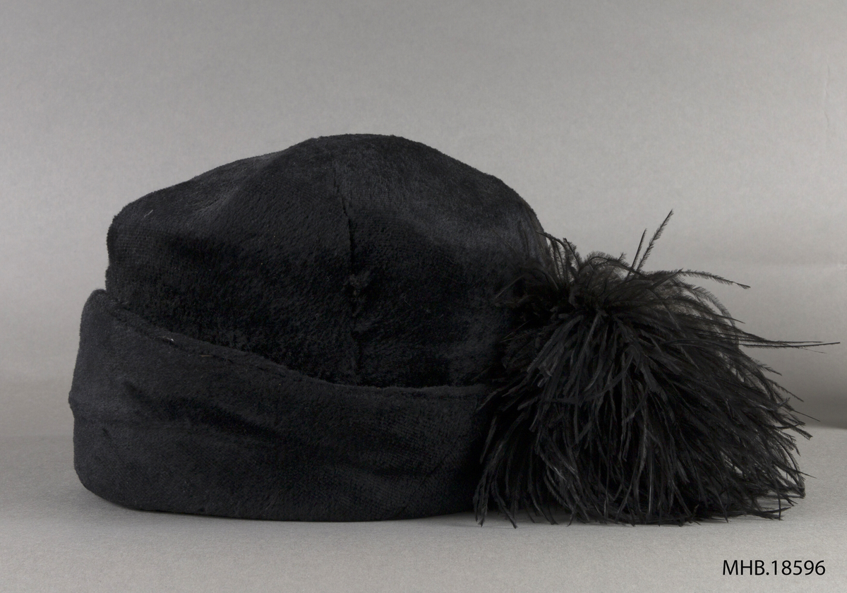 Hatt. Sort damehatt med fjær. Rund hatt med oppbrettet brem. Pull og brem i ett stykk. Bremmen brettes opp: Bredde foran og litt smaler bak på nakken, hvor fjærpynten er sydd på.