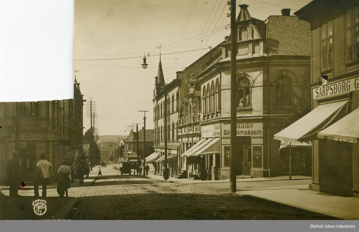 St. Marie gate i Sarpsborg sett mot torvet med Matstova midt i bildet. Baker Halvorsens butikk til venstre, til høyre Sarpsborg Glasmagasin og Raade Skotøimagasin, begge hjørnebygninger. Kiosk på torget. Bil og folk i gatene.
Postkort.