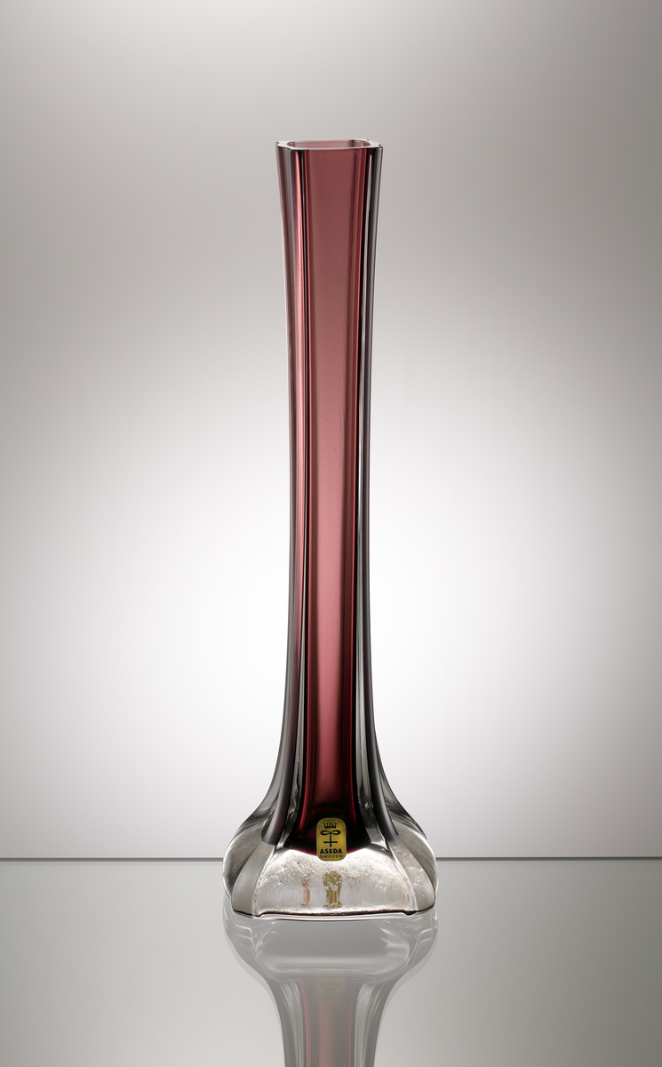 Hög, smal vas i vinrött underfång. Från den breda klackan strävar fyra stående linjer i relief.