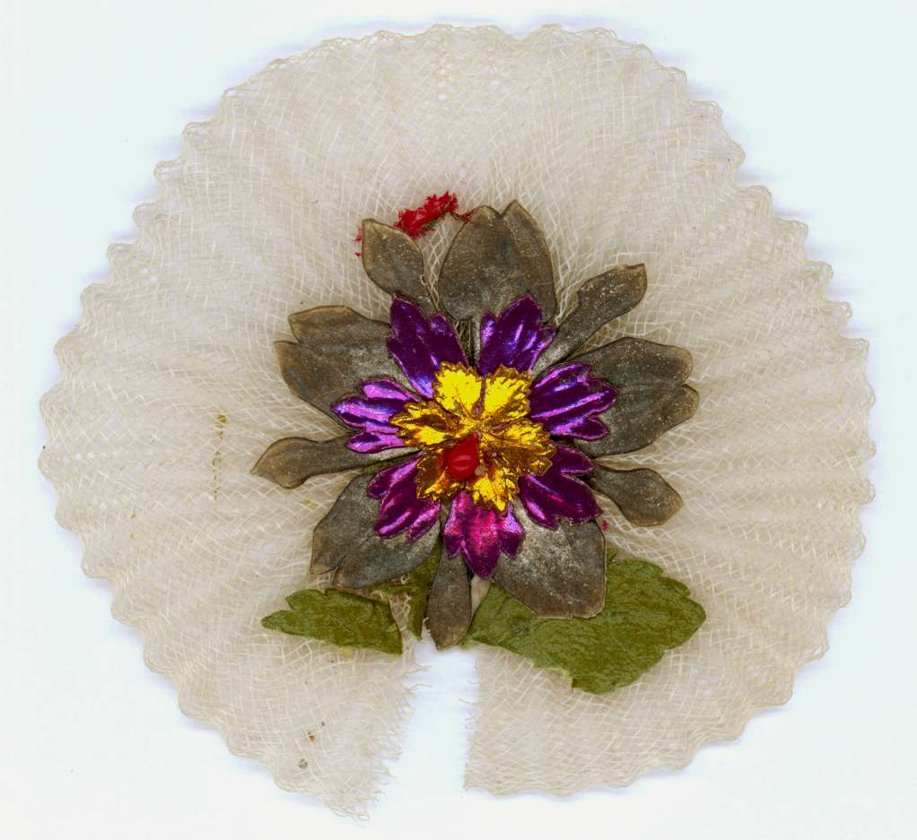 Runt kotiljongsmärke av vit tyll med påklistrad blomma av papper i grönt, grått, lila, guld och rött.