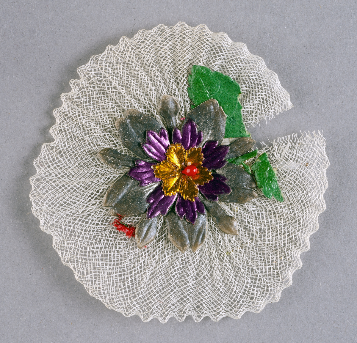 Runt kotiljongsmärke av vit tyll med påklistrad blomma av papper i grönt, grått, lila, guld och rött.