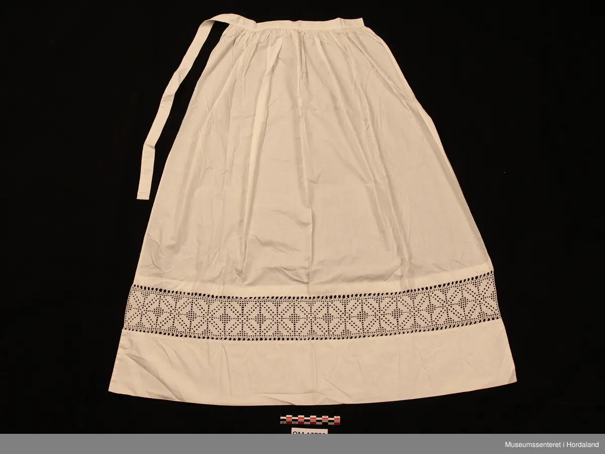Kvitt bundasforkle med hekla borde nede, innfelt i forkleet. Firkløverliknande motiv. Bommulslerretet som forkleet er sydd av er av usedvanleg tett og fin kvalitet (dunlerret). Oppe i livet er forkleet rynket. Det er sydd på linning som fortsetter rundt som forkleband.