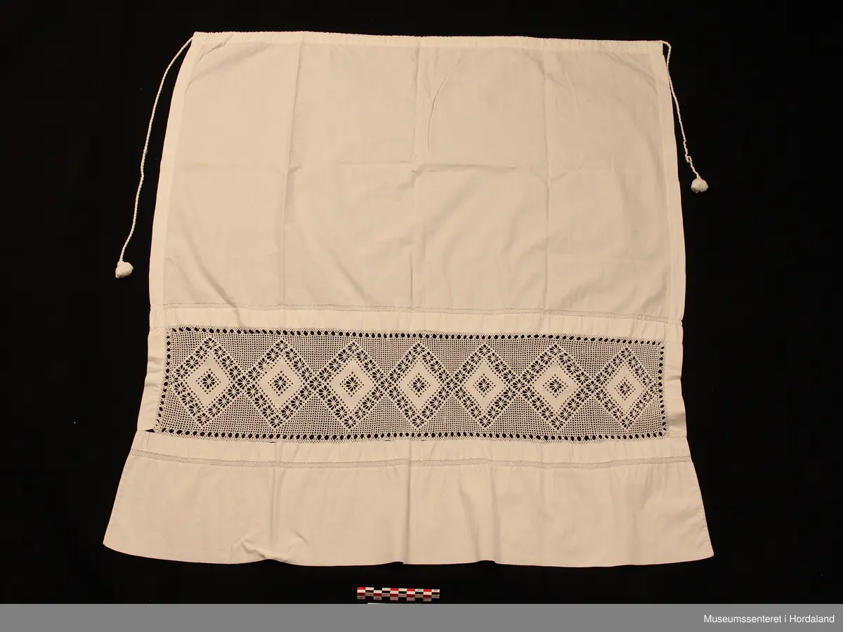 Kvitt bundasforkle med hekla  borde, innfelt i forkleet nede. Mønster inndelt i romber.På kvar side av borda er det ein smal saum med holfald. Oppe i livet er det sydd på ein løpegang, der det går ei tvinna snor igjennomtil å knyta bak, duskar i endene.