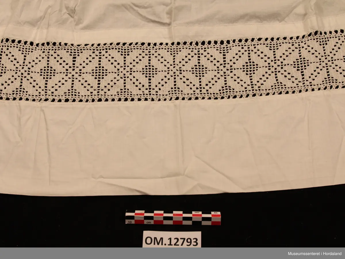 Kvitt bundasforkle med hekla borde nede, innfelt i forkleet. Firkløverliknande motiv. Bommulslerretet som forkleet er sydd av er av usedvanleg tett og fin kvalitet (dunlerret). Oppe i livet er forkleet rynket. Det er sydd på linning som fortsetter rundt som forkleband.