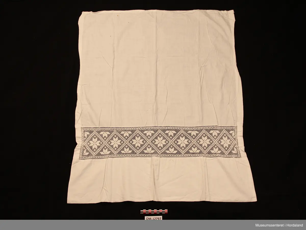 Kvitt bundasforkle med filert borde, innfelt nede. Motivet er åttebladroser. Oppe i livet er det bretta ned til ein løpegang, der snora mangler. Forklet er litt flekkete (rustflekker) og har nokre små hol.