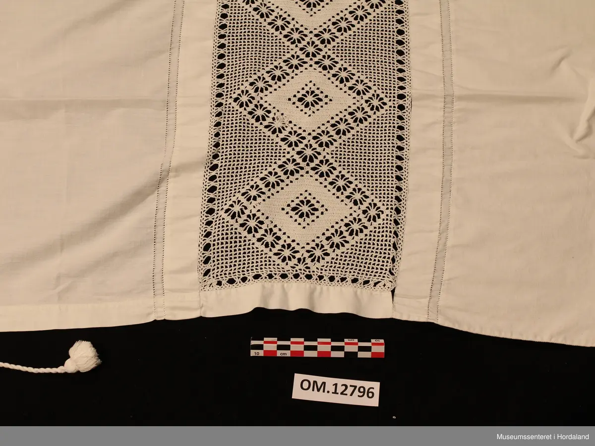 Kvitt bundasforkle med hekla  borde, innfelt i forkleet nede. Mønster inndelt i romber.På kvar side av borda er det ein smal saum med holfald. Oppe i livet er det sydd på ein løpegang, der det går ei tvinna snor igjennomtil å knyta bak, duskar i endene.