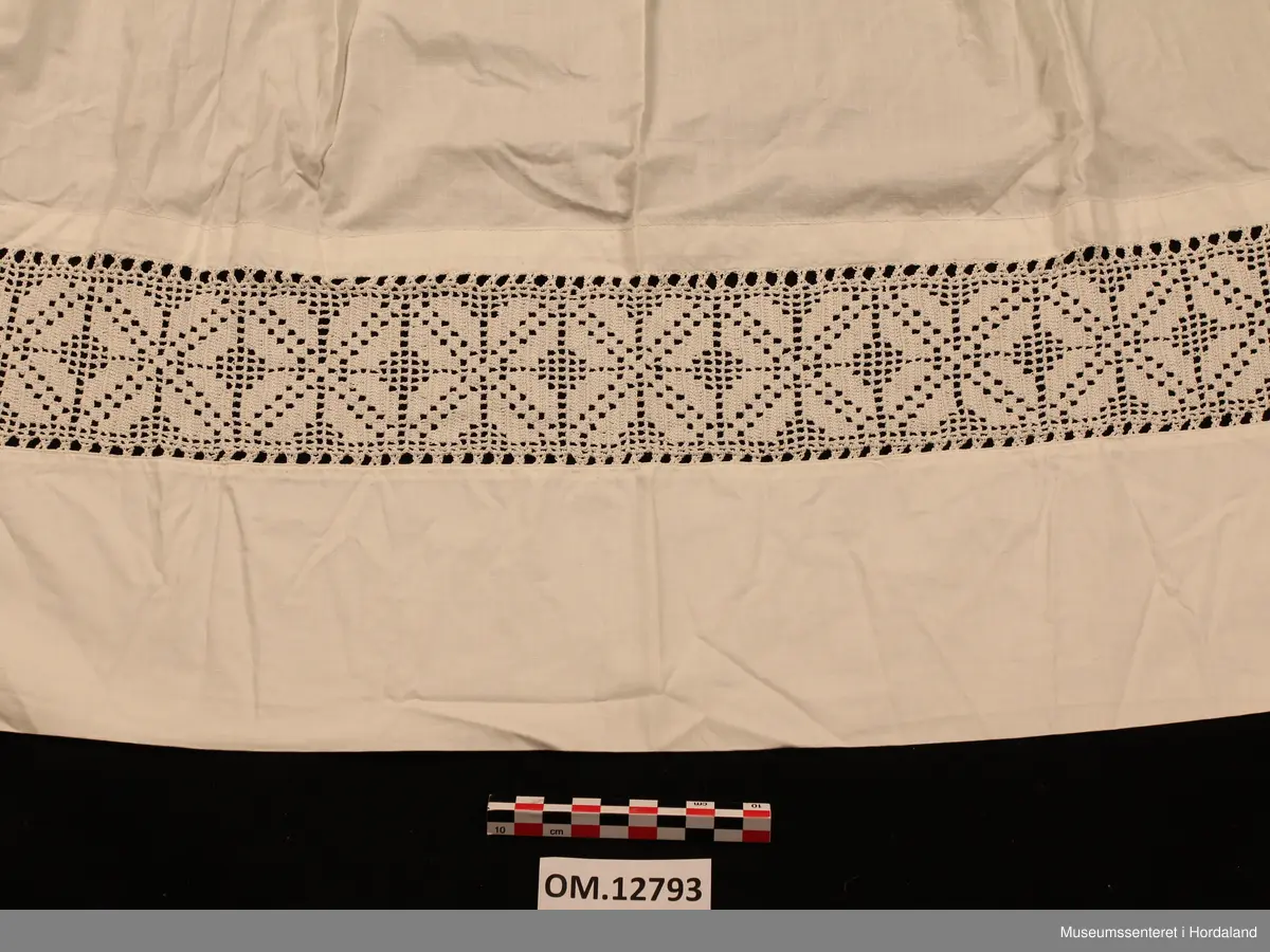 Kvitt bundasforkle med hekla borde nede, innfelt i forkleet. Firkløverliknande motiv. Bommulslerretet som forkleet er sydd av er av usedvanleg tett og fin kvalitet (dunlerret). Oppe i livet er forkleet rynket. Det er sydd på linning som fortsetter rundt som forkleband.