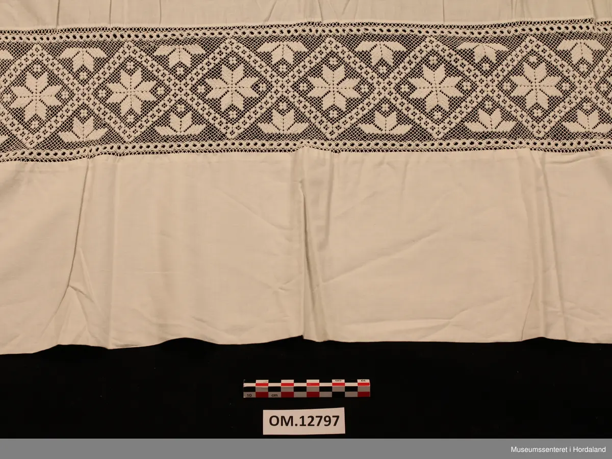 Kvitt bundasforkle med filert borde, innfelt nede. Motivet er åttebladroser. Oppe i livet er det bretta ned til ein løpegang, der snora mangler. Forklet er litt flekkete (rustflekker) og har nokre små hol.