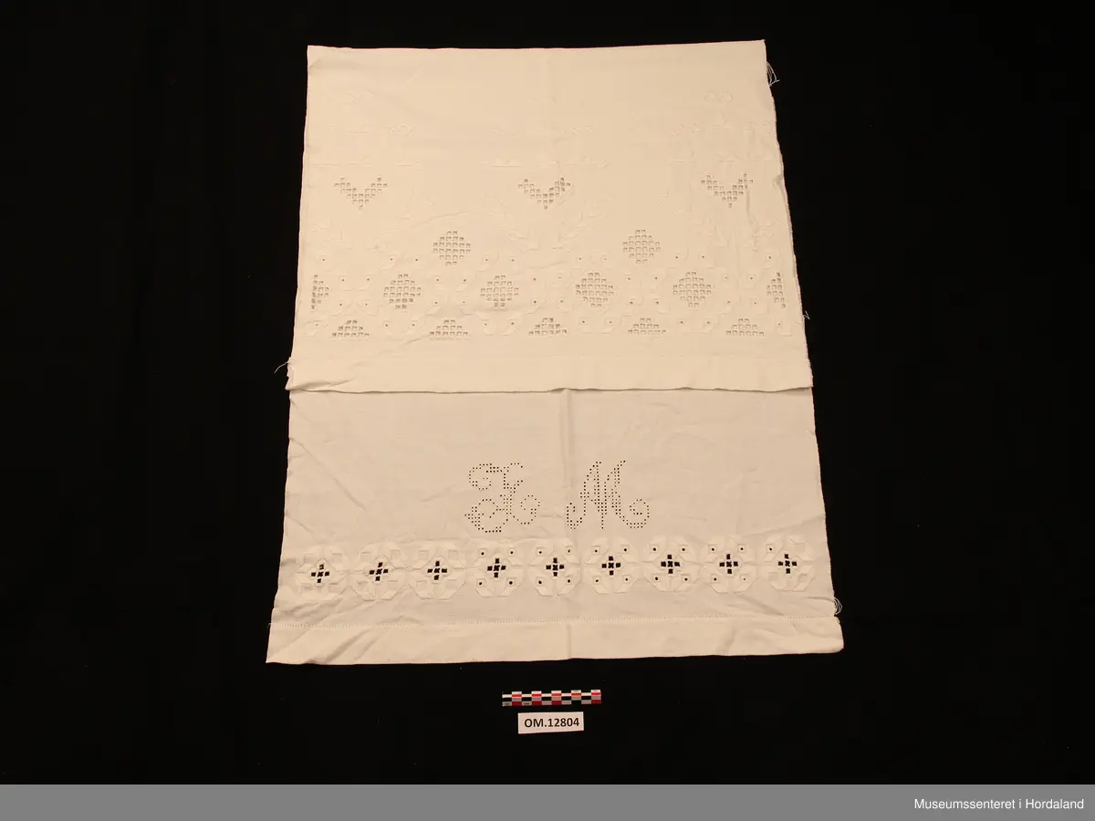 Rektangulært pyntehandkle med stormønstra, nokså fri hardangersaum (nokså grov kvitsaum - hardangersaumen grenser litt til plattsaum med primhol inni ). Sydd på javastoff. På det stykket som skal henge fram over stanga, er det ei pynteborde (plattsaum med primhol inni), samt eit monogram sydd av primhol - L M). Primhola i borda er ikkje ferdigsydde (slutta midt inni eit primhol). Utskurdsaumen og primhola er sydde i tynt hardtvinna bomullsgarn. Plattsaumen er sydd med perlegarn. Handkleet er falda med enkel holfald. Jarekant på begge langsider.