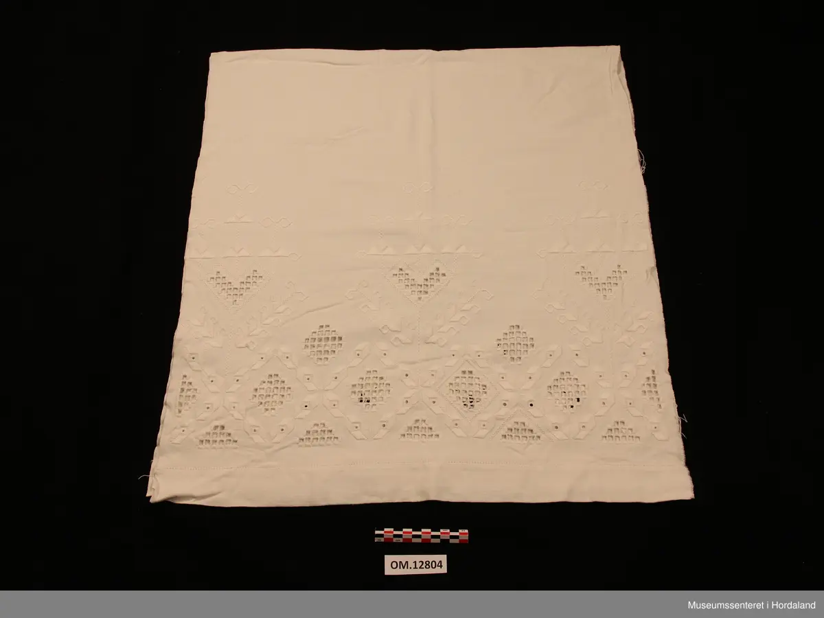 Rektangulært pyntehandkle med stormønstra, nokså fri hardangersaum (nokså grov kvitsaum - hardangersaumen grenser litt til plattsaum med primhol inni ). Sydd på javastoff. På det stykket som skal henge fram over stanga, er det ei pynteborde (plattsaum med primhol inni), samt eit monogram sydd av primhol - L M). Primhola i borda er ikkje ferdigsydde (slutta midt inni eit primhol). Utskurdsaumen og primhola er sydde i tynt hardtvinna bomullsgarn. Plattsaumen er sydd med perlegarn. Handkleet er falda med enkel holfald. Jarekant på begge langsider.