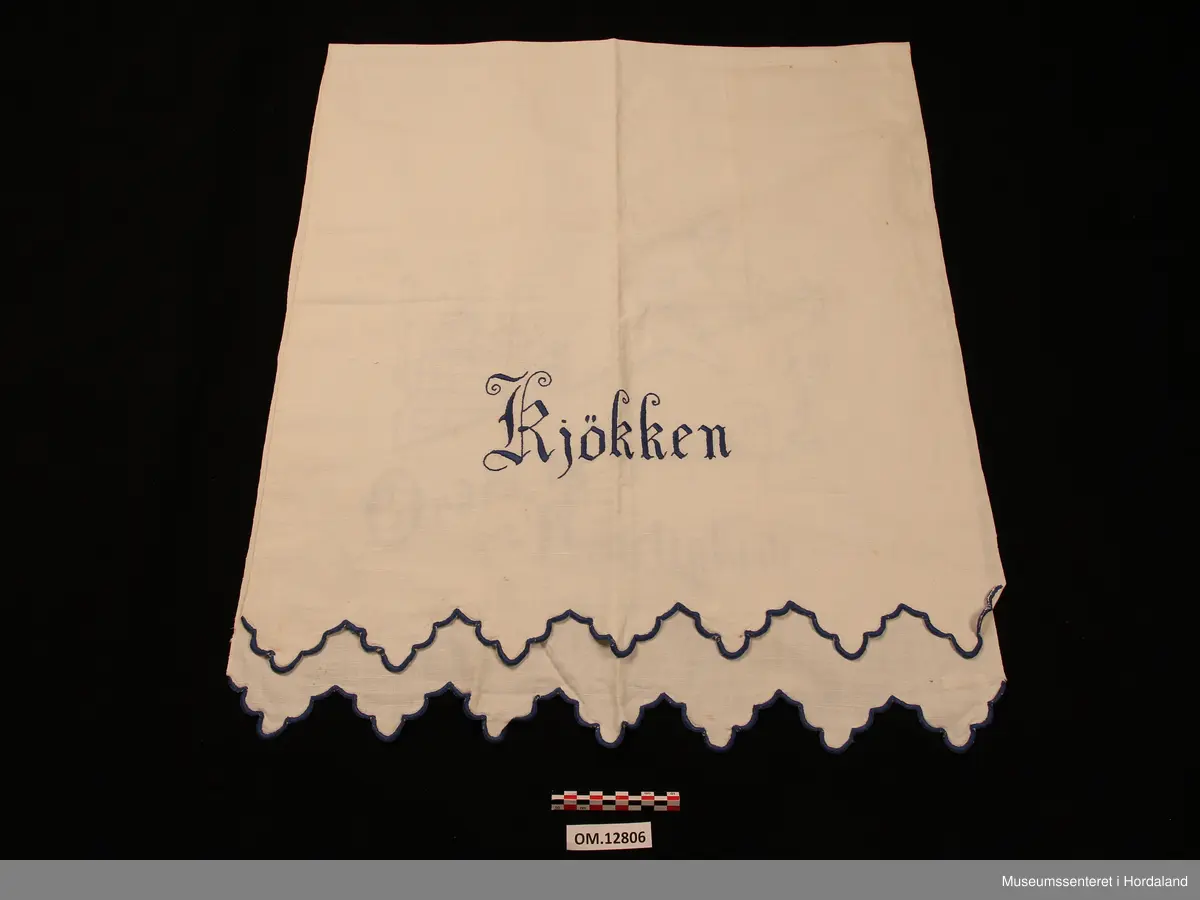 Rektangulært pyntehandkle med broderi i stilkesting (påteikna mønster).  Motivet er ei dame som står ved ein ovn med to pannar oppå. Nede er det tekst, der det står: Orden og renslighed. På den delen som skal hanga over stanga står det : Kjøkken (alt brodert i plattsaum). Avluttinga på begge endane er bodert i tungesting, klipt i ein tungekant (brodert først og klipt etterpå). Stoffet er av lin og det er jarekantar på begge langsider. Broderigarnet er av bomull.