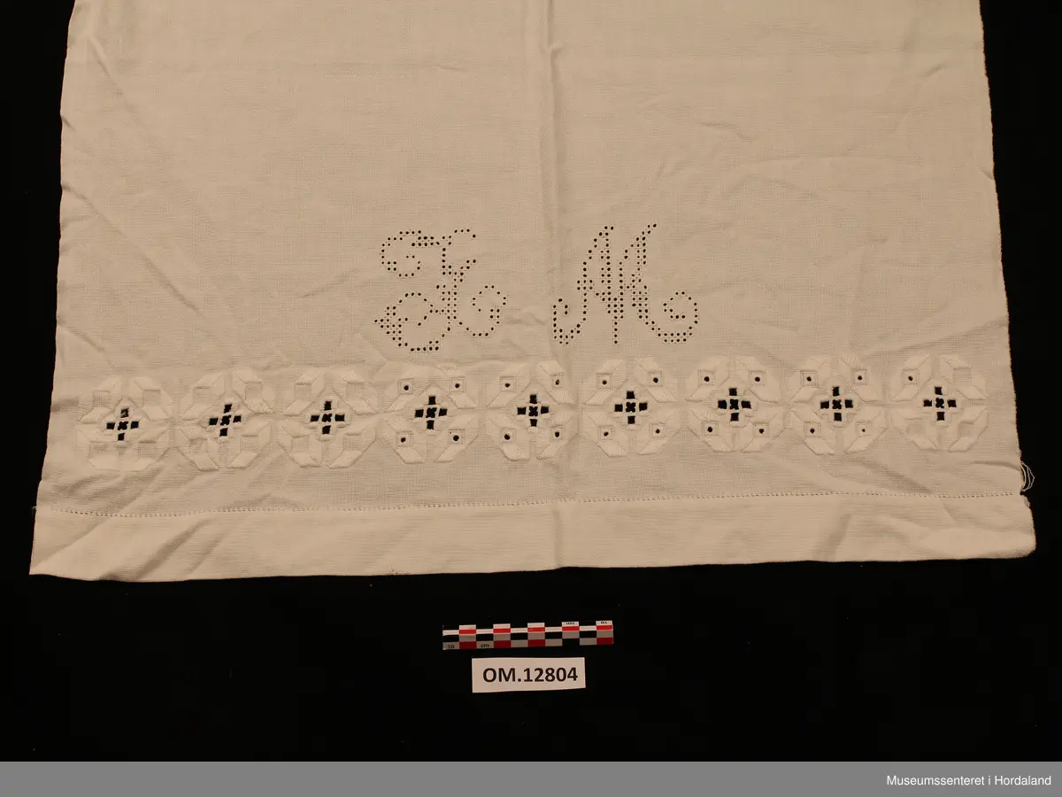 Rektangulært pyntehandkle med stormønstra, nokså fri hardangersaum (nokså grov kvitsaum - hardangersaumen grenser litt til plattsaum med primhol inni ). Sydd på javastoff. På det stykket som skal henge fram over stanga, er det ei pynteborde (plattsaum med primhol inni), samt eit monogram sydd av primhol - L M). Primhola i borda er ikkje ferdigsydde (slutta midt inni eit primhol). Utskurdsaumen og primhola er sydde i tynt hardtvinna bomullsgarn. Plattsaumen er sydd med perlegarn. Handkleet er falda med enkel holfald. Jarekant på begge langsider.