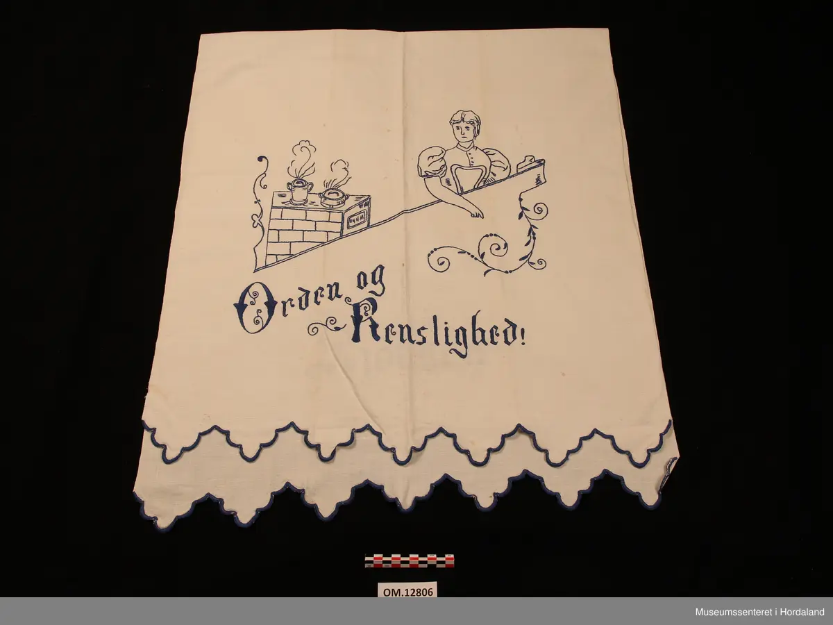 Rektangulært pyntehandkle med broderi i stilkesting (påteikna mønster).  Motivet er ei dame som står ved ein ovn med to pannar oppå. Nede er det tekst, der det står: Orden og renslighed. På den delen som skal hanga over stanga står det : Kjøkken (alt brodert i plattsaum). Avluttinga på begge endane er bodert i tungesting, klipt i ein tungekant (brodert først og klipt etterpå). Stoffet er av lin og det er jarekantar på begge langsider. Broderigarnet er av bomull.