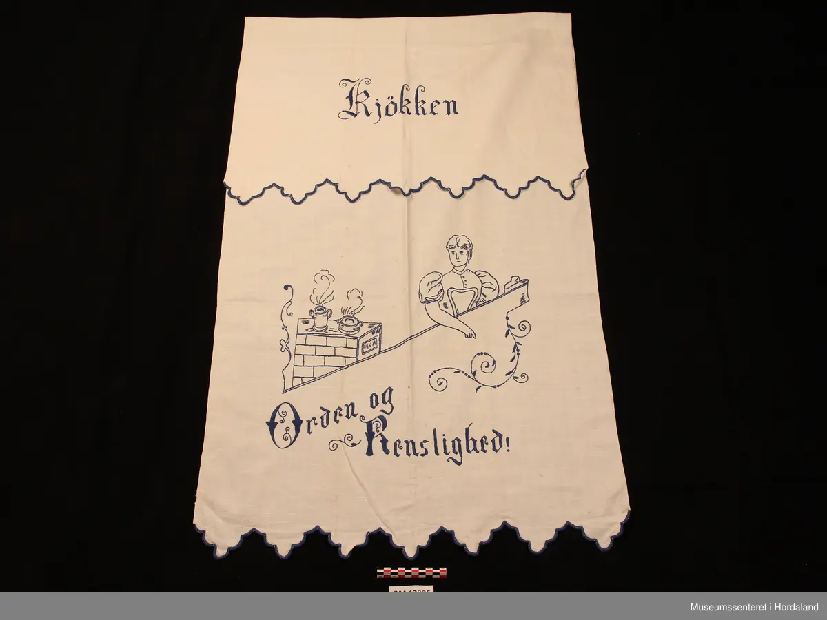 Rektangulært pyntehandkle med broderi i stilkesting (påteikna mønster).  Motivet er ei dame som står ved ein ovn med to pannar oppå. Nede er det tekst, der det står: Orden og renslighed. På den delen som skal hanga over stanga står det : Kjøkken (alt brodert i plattsaum). Avluttinga på begge endane er bodert i tungesting, klipt i ein tungekant (brodert først og klipt etterpå). Stoffet er av lin og det er jarekantar på begge langsider. Broderigarnet er av bomull.