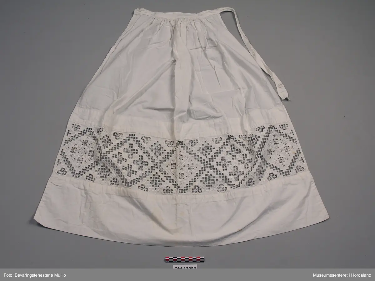 Kvitt bomullsforkle med brei hardangersaumbord innfelt nede. Nedre kanten er dobbel. Rynking ved linninga. Linning har tamp som går rundt livet. Knapp og knapphol. Forkledet er lagt inn i sidene. Nokre gule flekkar nedst.