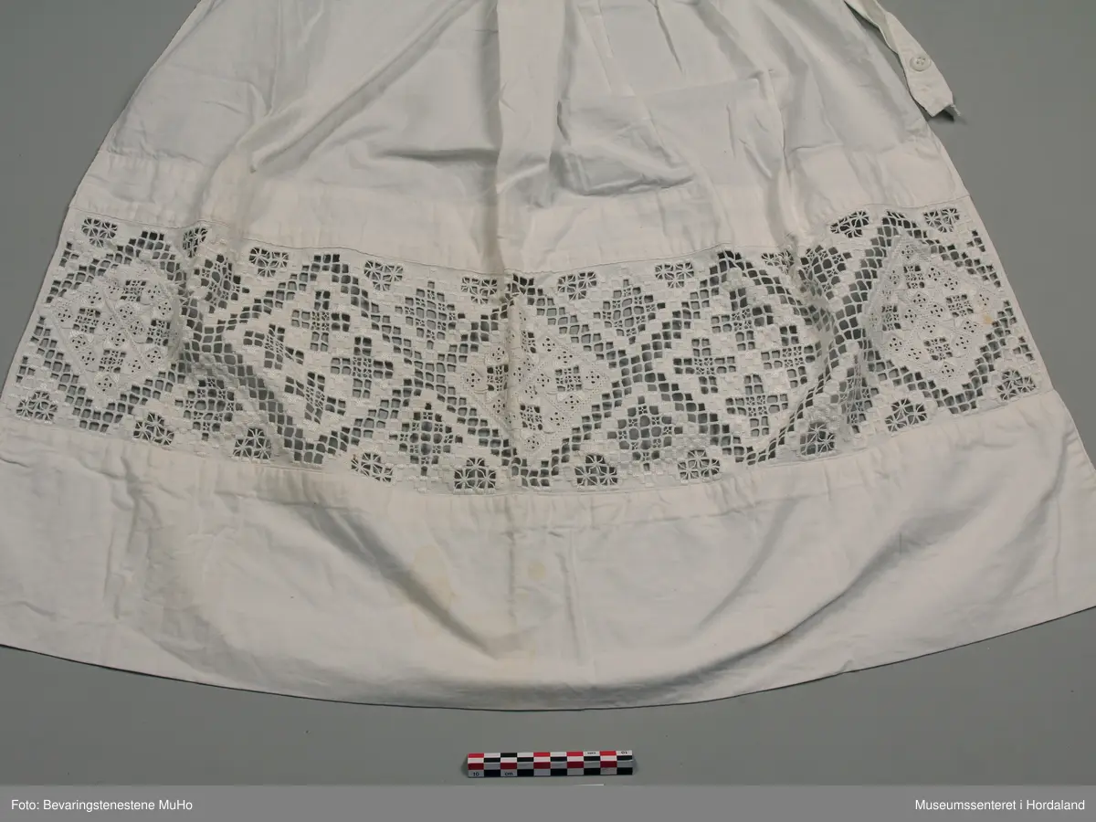 Kvitt bomullsforkle med brei hardangersaumbord innfelt nede. Nedre kanten er dobbel. Rynking ved linninga. Linning har tamp som går rundt livet. Knapp og knapphol. Forkledet er lagt inn i sidene. Nokre gule flekkar nedst.