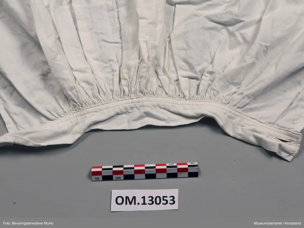 Kvitt bomullsforkle med brei hardangersaumbord innfelt nede. Nedre kanten er dobbel. Rynking ved linninga. Linning har tamp som går rundt livet. Knapp og knapphol. Forkledet er lagt inn i sidene. Nokre gule flekkar nedst.