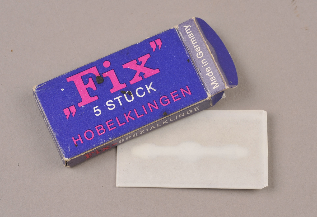 Pappask med lok. Asken har to rom. I asken er det fingerbeskyttelse, nålepresserar og høvelblad.