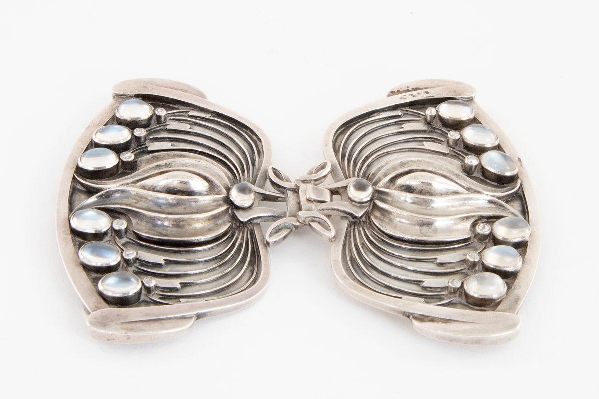 Todelt beltespenne i støpt og siselert sølv. De to spennedelene har en blomsterlignende form, og er dekorert med små diamanter og månesteiner. Disse er montert i gjennombrutte partier av metallet. Det er oppgitt at spennen opprinnelig har vært oksidert til en mørk gråblå tone, denne er nå borte, sølvet er blankt.