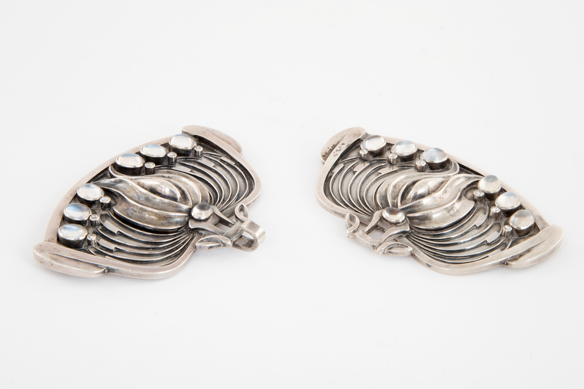 Todelt beltespenne i støpt og siselert sølv. De to spennedelene har en blomsterlignende form, og er dekorert med små diamanter og månesteiner. Disse er montert i gjennombrutte partier av metallet. Det er oppgitt at spennen opprinnelig har vært oksidert til en mørk gråblå tone, denne er nå borte, sølvet er blankt.