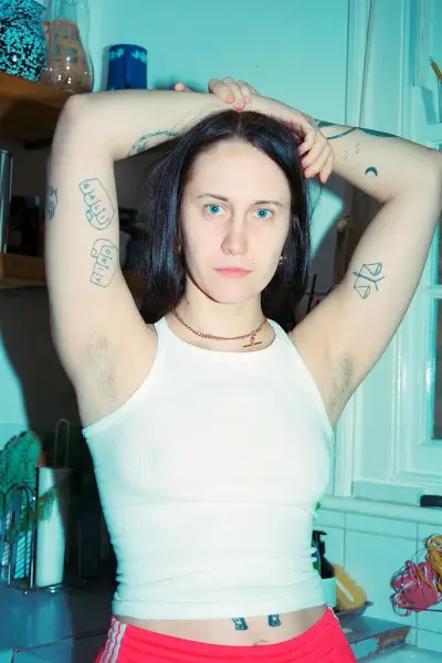 En kvinne står med armene over hodet. På armene har hun tatoveringer, hun har hvit trøye og røde bukser.