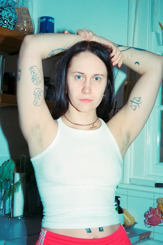 En kvinne står med armene over hodet. På armene har hun tatoveringer, hun har hvit trøye og røde bukser.