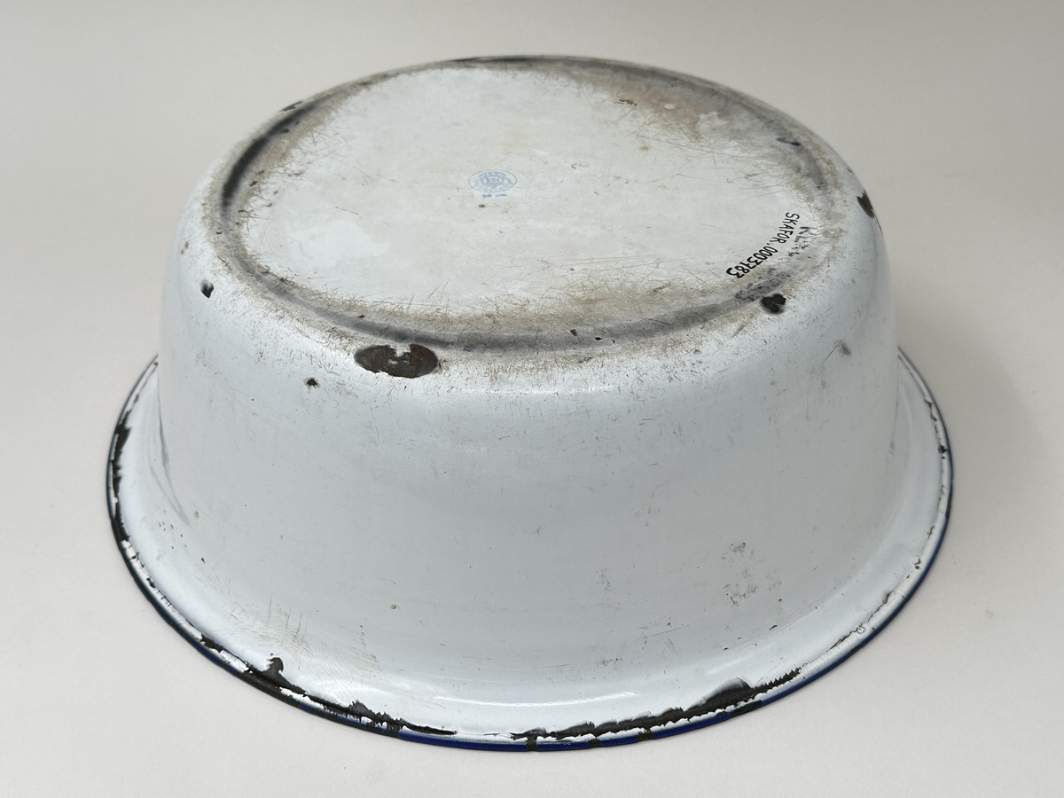 Skål, av emaljerad metall. Vit emalj med rullad blå kant. Stämpel i botten. Emaljen kantstött.