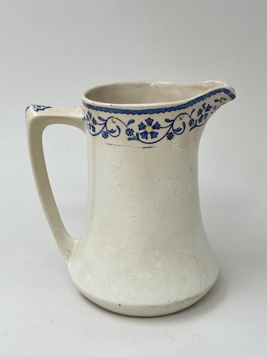 Tillbringare, fabriksgjord av krämfärgat flintgods, kromtryckt under glasyren. Modell med konkav sida, rakt handtag och pip. Dekoren i två blå toner samt gult är placerad längs överkanten som en blomsterslinga och på handtaget. Märkt i botten med blått på det vita: ankare - band med GUSTAF - band med Gustafsberg, några svårtydda stämplar i godset.
Lätt krackelerad över hela ytan. Spricka i överkanten samt på insidan vid handtagsfästet. 