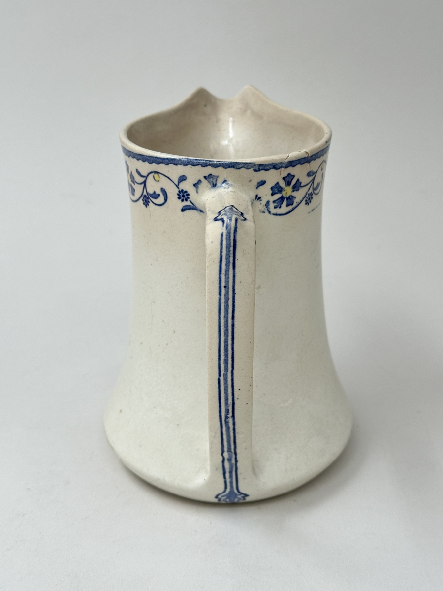 Tillbringare, fabriksgjord av krämfärgat flintgods, kromtryckt under glasyren. Modell med konkav sida, rakt handtag och pip. Dekoren i två blå toner samt gult är placerad längs överkanten som en blomsterslinga och på handtaget. Märkt i botten med blått på det vita: ankare - band med GUSTAF - band med Gustafsberg, några svårtydda stämplar i godset.
Lätt krackelerad över hela ytan. Spricka i överkanten samt på insidan vid handtagsfästet. 