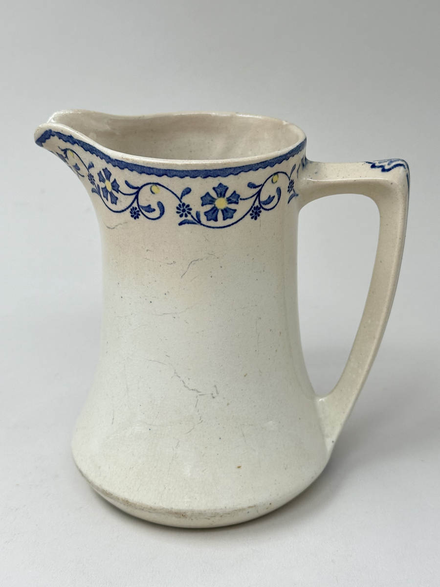 Tillbringare, fabriksgjord av krämfärgat flintgods, kromtryckt under glasyren. Modell med konkav sida, rakt handtag och pip. Dekoren i två blå toner samt gult är placerad längs överkanten som en blomsterslinga och på handtaget. Märkt i botten med blått på det vita: ankare - band med GUSTAF - band med Gustafsberg, några svårtydda stämplar i godset.
Lätt krackelerad över hela ytan. Spricka i överkanten samt på insidan vid handtagsfästet. 