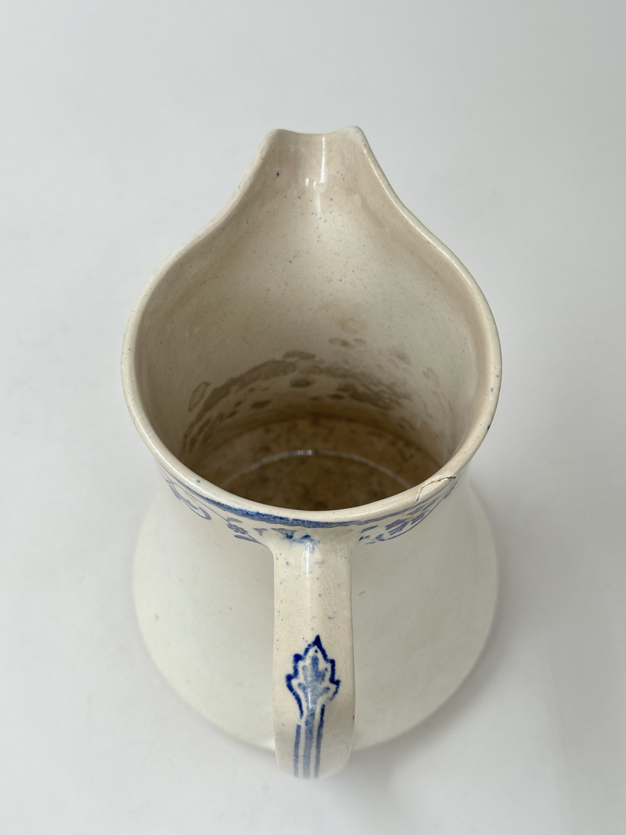 Tillbringare, fabriksgjord av krämfärgat flintgods, kromtryckt under glasyren. Modell med konkav sida, rakt handtag och pip. Dekoren i två blå toner samt gult är placerad längs överkanten som en blomsterslinga och på handtaget. Märkt i botten med blått på det vita: ankare - band med GUSTAF - band med Gustafsberg, några svårtydda stämplar i godset.
Lätt krackelerad över hela ytan. Spricka i överkanten samt på insidan vid handtagsfästet. 