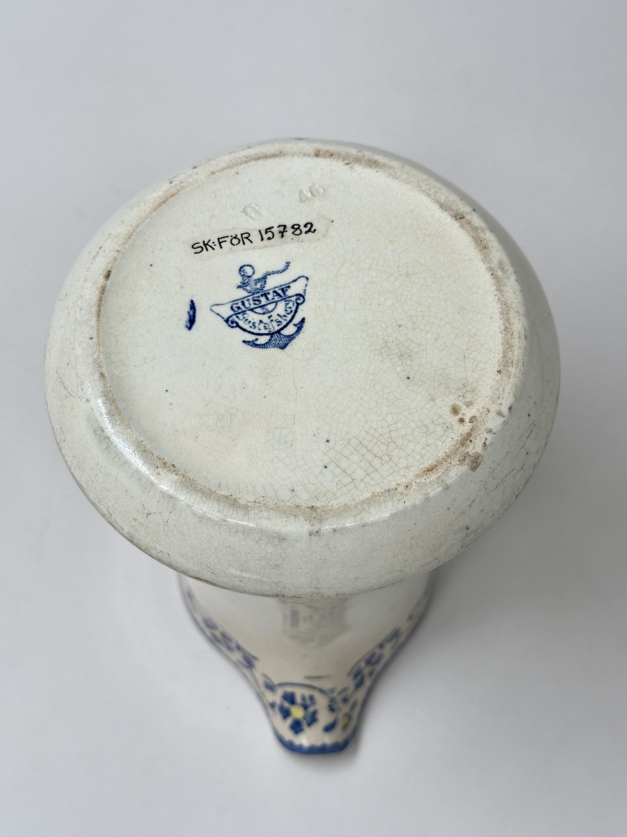 Tillbringare, fabriksgjord av krämfärgat flintgods, kromtryckt under glasyren. Modell med konkav sida, rakt handtag och pip. Dekoren i två blå toner samt gult är placerad längs överkanten som en blomsterslinga och på handtaget. Märkt i botten med blått på det vita: ankare - band med GUSTAF - band med Gustafsberg, några svårtydda stämplar i godset.
Lätt krackelerad över hela ytan. Spricka i överkanten samt på insidan vid handtagsfästet. 