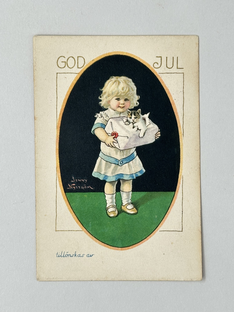 Julkort i vit kartong. Oval bild: Svart vägg, grönt golv. Blont barn i vitt och ljusblått håller paket ur vars inre en katt tittar ut. Guldtext: GOD JUL. Frankerat med rött femöresfrimärke med lejon. Stämplat 25/12 1923.