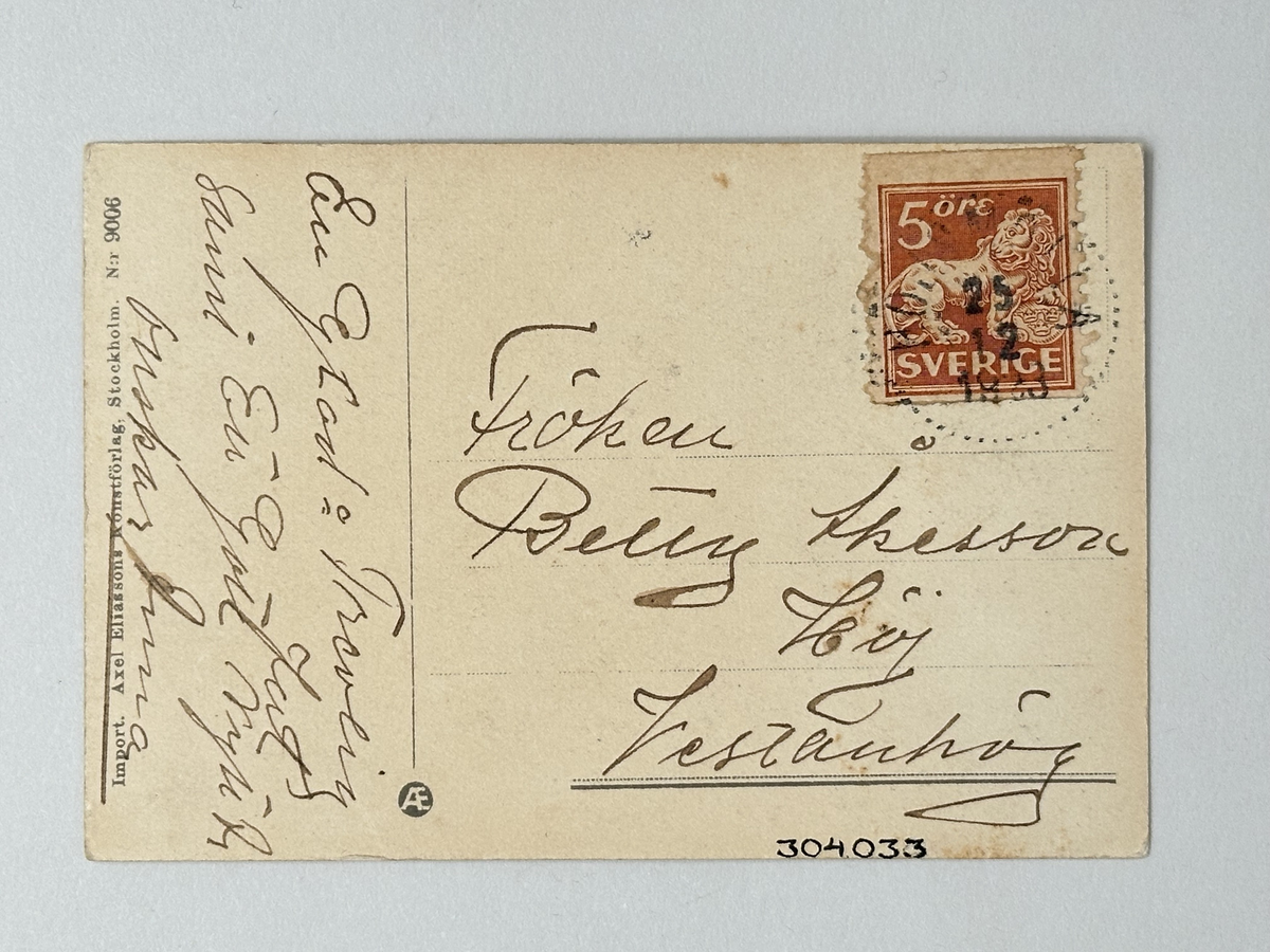 Julkort i vit kartong. Oval bild: Svart vägg, grönt golv. Blont barn i vitt och ljusblått håller paket ur vars inre en katt tittar ut. Guldtext: GOD JUL. Frankerat med rött femöresfrimärke med lejon. Stämplat 25/12 1923.