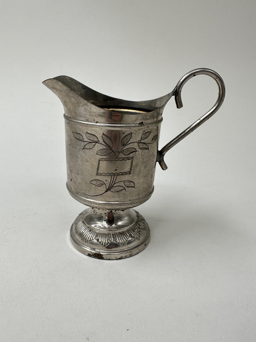 Gräddsnipa i nickel. Cicelerad dekor i form av bladrankor och parallellogram. Lodgrepe. Hög fot med spetsig dekor i reliefränder.