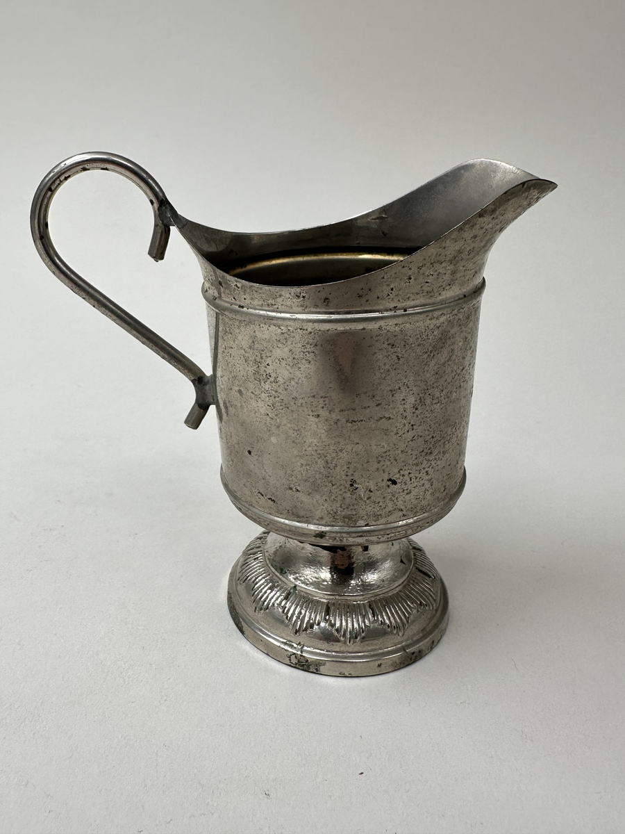 Gräddsnipa i nickel. Cicelerad dekor i form av bladrankor och parallellogram. Lodgrepe. Hög fot med spetsig dekor i reliefränder.