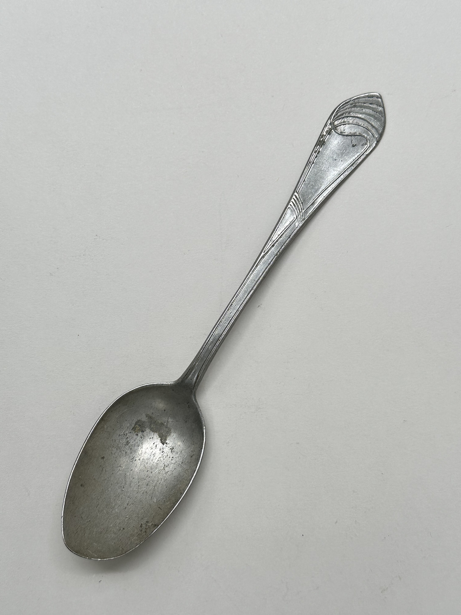 Sked i aluminiumlegering. Dekor av slingor i jugendstil på skaftet. Oregelbunden form på bladet. 