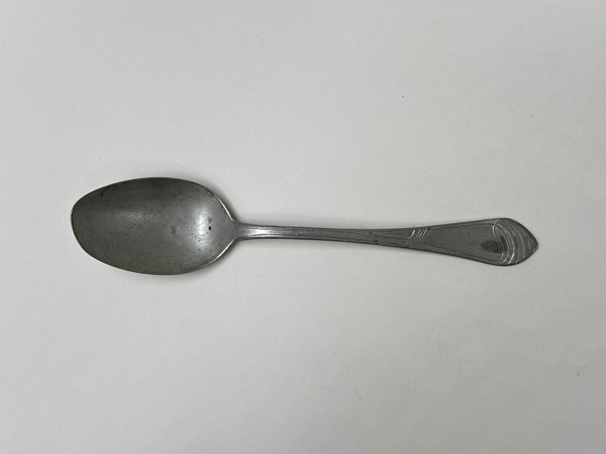 Sked i aluminiumlegering. Dekor av slingor i jugendstil på skaftet. Oregelbunden form på bladet. 