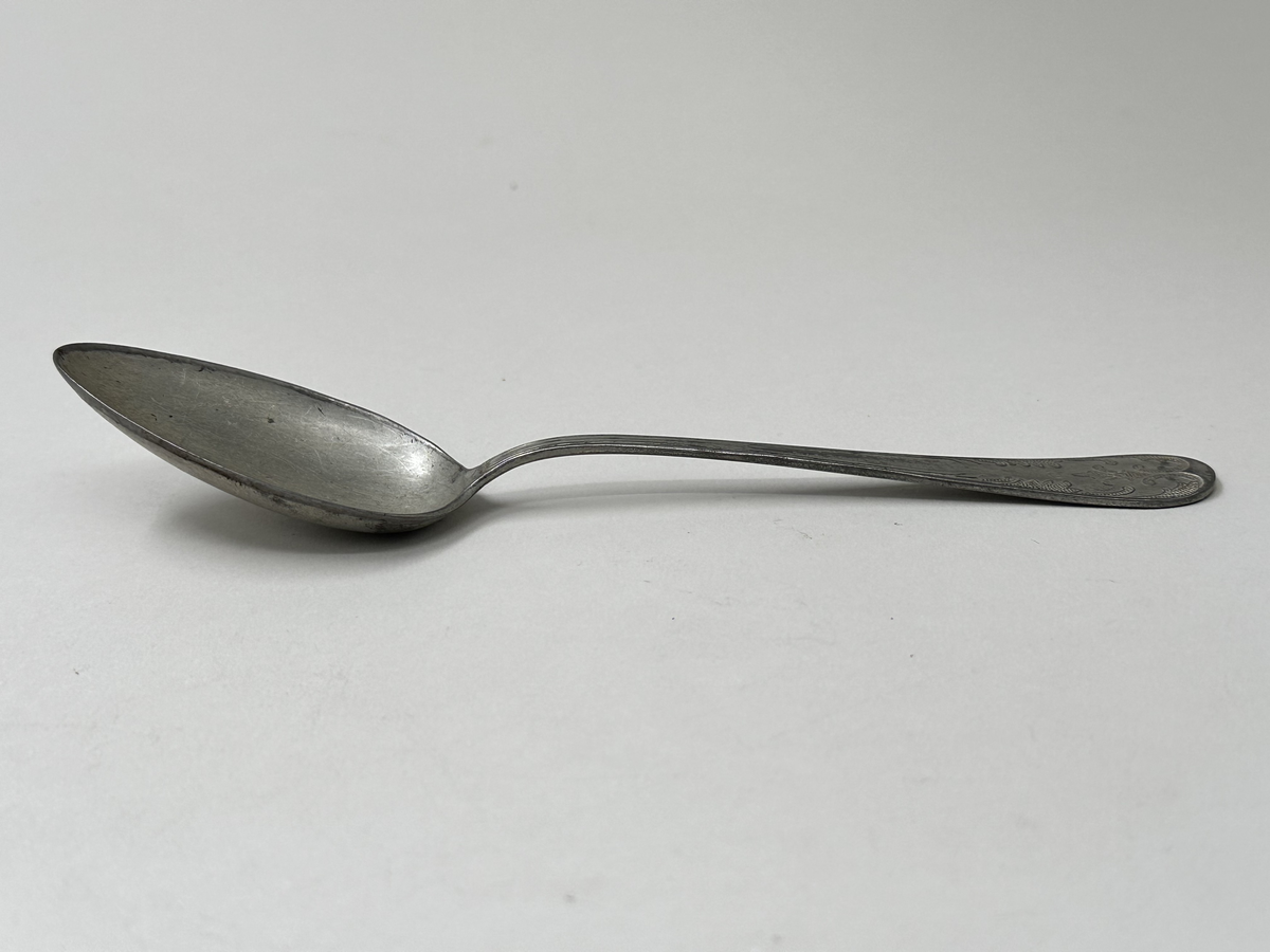 Sked, av vit metallegering. Skaftets ovansida dekorerad med gammalfranskt mönster. Undersidan stämplad med "Alpa Rostskydd Imp."