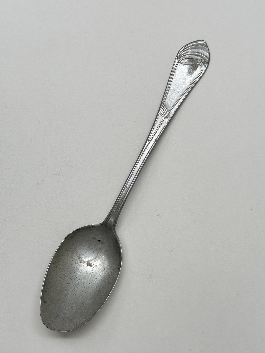 Sked i aluminiumlegering. Dekor av slingor i jugendstil på skaftet. Oregelbunden form på bladet. 