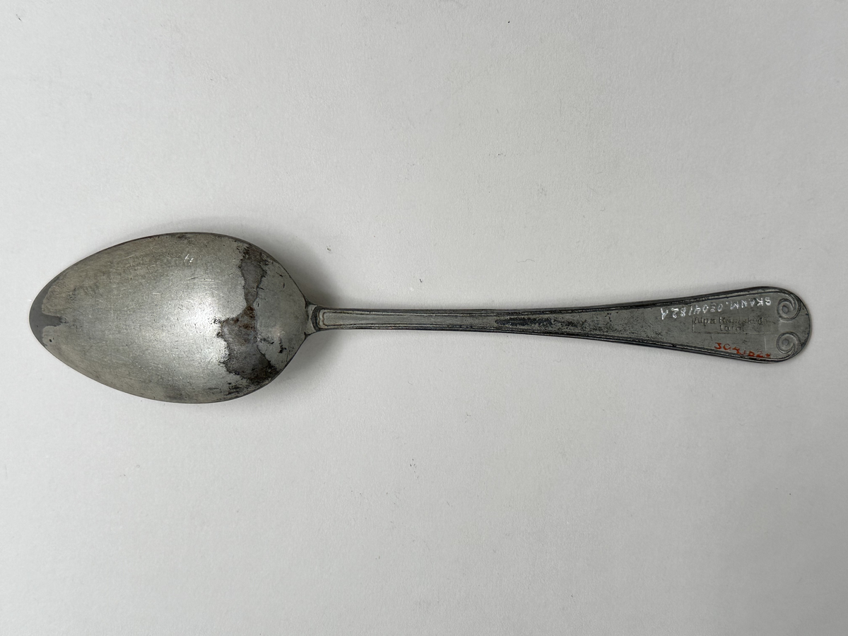 Sked, av vit metallegering. Skaftets ovansida dekorerad med gammalfranskt mönster. Undersidan stämplad med "Alpa Rostskydd Imp."