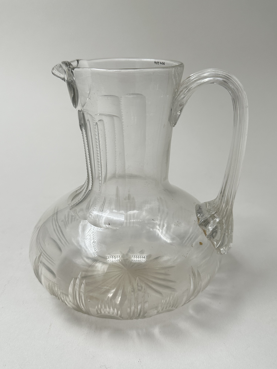 Tillbringare i glas. Munblåst. Bred buk, hög hals med snip samt lodgrepe. Slipad dekor; stjärna i botten samt ovaler och streck längs buken. Halsen med sex stycken stående platta slipningar; de två mittersta något kortare. 