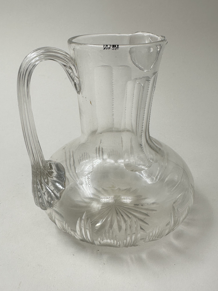 Tillbringare i glas. Munblåst. Bred buk, hög hals med snip samt lodgrepe. Slipad dekor; stjärna i botten samt ovaler och streck längs buken. Halsen med sex stycken stående platta slipningar; de två mittersta något kortare. 