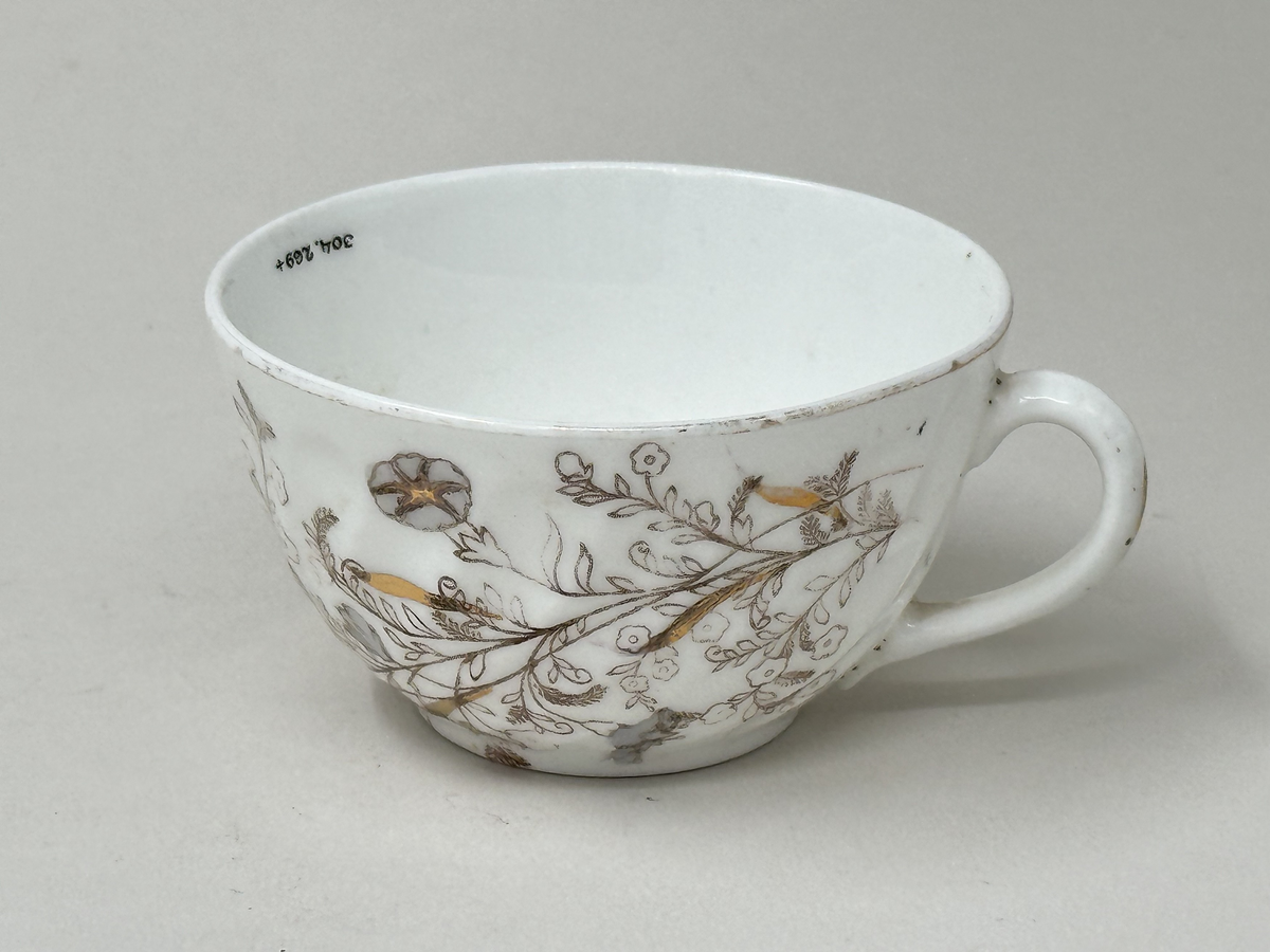 Koppar i vitt fältspatsporslin med tryckt blomdekor i brunt, guld och grått. Guldrand längs mynning och öra (delvis avskavt). Vriden godronnering runtom.